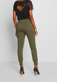 Zwarte top met kantdetail op de rug, gecombineerd met olijfgroene joggingbroek met een zijzak en elastische boorden, gedragen met zwarte hakken.