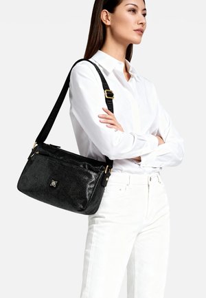 Borsa nera testurizzata con logo dorato, tracolla regolabile e scomparti con cerniera, tenuta da una persona con camicia bianca con bottoni.