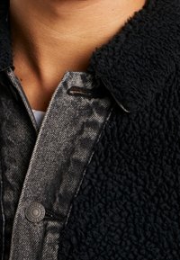 Musta pörröinen fleece-kaulus harmaan farkkupaidan päällä, jossa on näyttävä nappi kaiverretulla tekstillä ja tekstuurilla oleva kangassynergi.