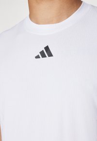 Bela atletska majica z rebrasto teksturo in črnim Adidasovim logotipom na prsih. Ima okrogel izrez in obliko, ki se prilega.