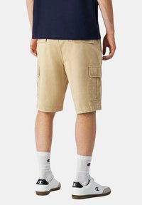 Pantaloni cargo beige con due tasche laterali, taglio dritto e orlo cucito, indossati con sneakers bianche e calze bianche a coste.