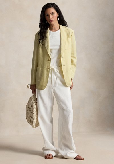 Femme portant un blazer jaune pâle, un t-shirt blanc, un pantalon large blanc, des sandales marron, tenant un sac fourre-tout tissé beige devant un fond neutre.