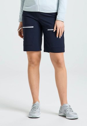 Un modelo lleva unos pantalones cortos cargo azul marino hasta la rodilla, con bolsillos con cremallera, una chaqueta blanca y zapatillas deportivas gris claro.
