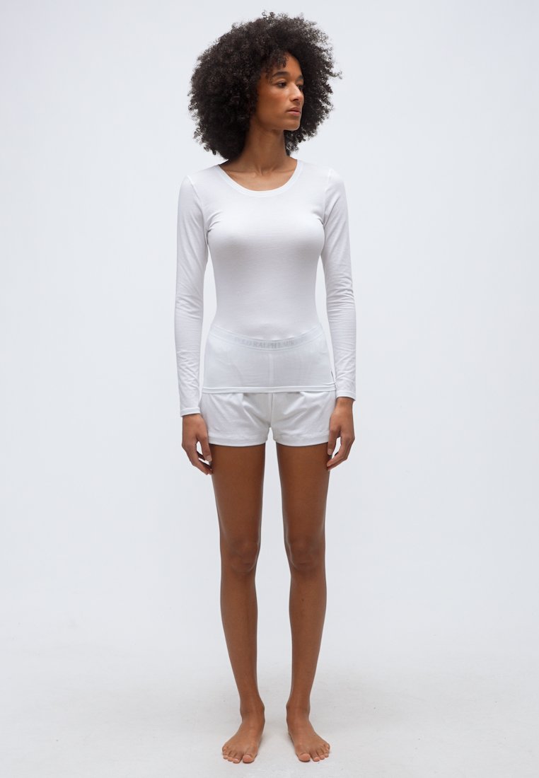 Femme aux cheveux bouclés, pieds nus, portant un haut blanc à manches longues ajusté et un short blanc, debout devant un fond uni clair.