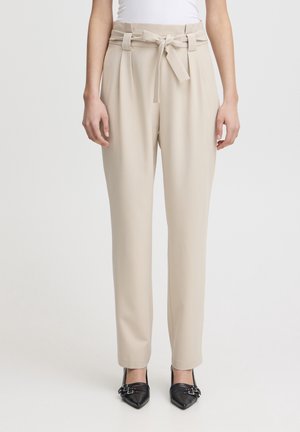 Beige paperbag-bukser med højt liv og bundet bælte, båret med sorte spidse hæle og en hvid top, stående mod en ensfarvet baggrund.