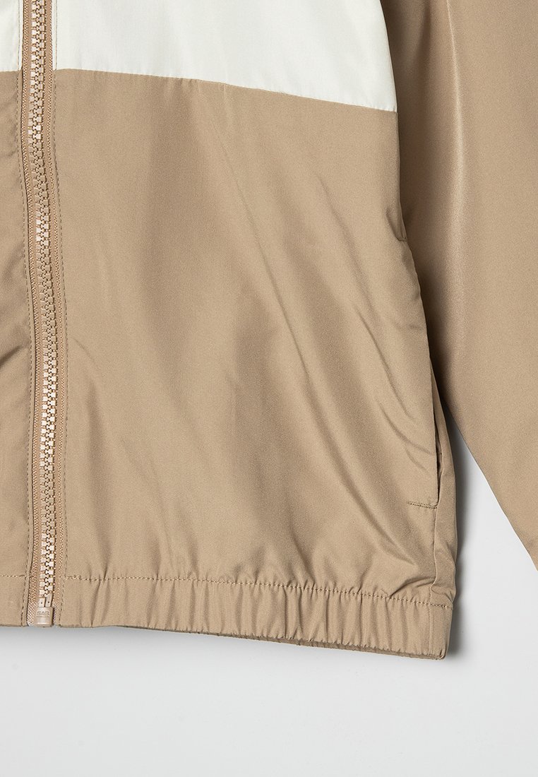 Veste coupe-vent beige et blanche avec une fermeture éclair intégrale, des poignets élastiques et un ourlet élastiqué. Tissu lisse et léger avec des poches latérales.