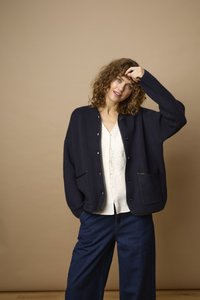 Femme aux cheveux bouclés portant un cardigan foncé sur une blouse blanche et un pantalon large bleu, touchant ses cheveux, debout devant un fond beige uni.