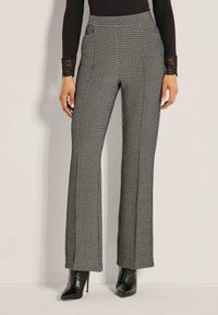 Frau, die hochtaillierte schwarze und weiße gemusterte weit geschnittene Hose mit schwarzen spitzen High Heels trägt, steht vor einem einfarbigen Hintergrund.