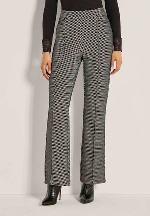 Vrouw in een hoge taille met zwart-wit gestreepte wijde broek met zwarte spitse hoge hakken, staande tegen een effen achtergrond.