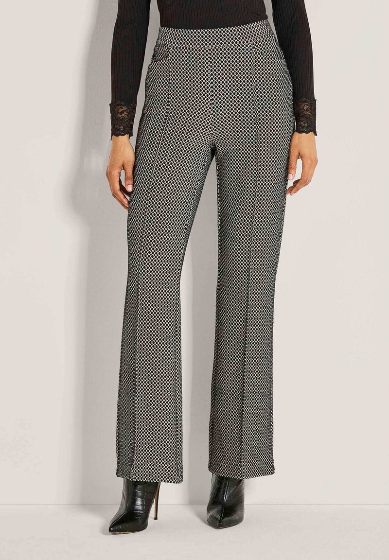 Frau, die hochtaillierte schwarze und weiße gemusterte weit geschnittene Hose mit schwarzen spitzen High Heels trägt, steht vor einem einfarbigen Hintergrund.