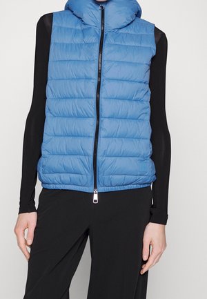 Personne portant un gilet matelassé bleu sans manches fermé par une fermeture éclair sur un haut noir à manches longues et un pantalon noir, sur un fond uni.