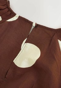 Hnedá tkanina s veľkými krémovými bodkami, ktorá obsahuje volánikový výstrih a detail s otvorom. Jemná textúra so subtílnym leskom.