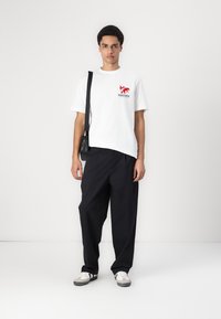 Hvid t-shirt med et rødt grafisk design og tekst, parret med sorte løstsiddende bukser og grå sneakers. En simpel sort skuldertaske medfølger.