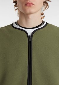 Giacca in fleece verde oliva con collo rotondo, zip nera e dettagli neri a contrasto. Presenta una texture morbida e un design minimalista.