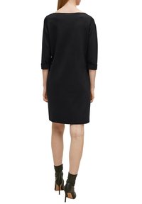 Robe noire arrivant au genou avec des manches trois-quarts, présentant un large décolleté. Portée avec des bottines à talons noires, mettant en valeur une texture lisse.