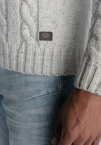 Suéter de punto gris claro con patrón de trenzas y puños acanalados. Presenta una pequeña etiqueta oscura en la parte frontal y se combina con jeans de color azul claro.