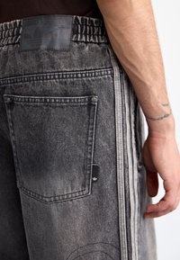 Close-up van een persoon in vervaagde zwarte Adidas-jeans met een elastische tailleband, een achterzak, drievoudige zijstrepen en een zichtbare getatoeëerde onderarm.