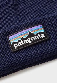 Jūrinės spalvos megztas kepuraitė su tekstūruotu raštu. Papuošta juodu siuvinėjimu su spalvingu kalnų dizainu ir "Patagonia" logotipu.