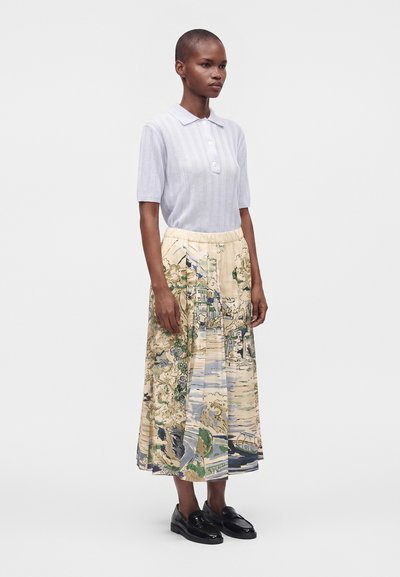 Femme portant un polo bleu clair à manches courtes, une jupe beige avec des imprimés colorés de paysages, et des mocassins noirs, debout devant un fond blanc.