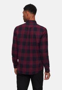 Uomo di spalle che indossa una camicia a quadri bordeaux e nera a maniche lunghe con pantaloni neri su uno sfondo grigio chiaro.