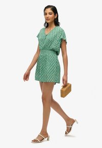 Vestido corto verde con estampado, escote en V, mangas cortas y cintura elástica, combinado con sandalias de tacón blancas y un bolso de caja de mimbre.