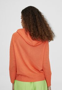 Hooded sweatshirt in bright orange with ribbed detailing on sleeves, soft texture, and relaxed fit, paired with green pants.