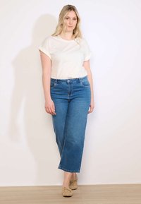 Heledate juustega naine valges lühikeste varrukatega särkis, sinistes laiade säärtega teksapükstes ja beežides mokassiinides, seisab ühevärvse valge seina taustal.