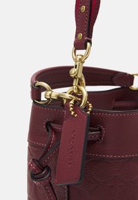 Borsa a secchiello in pelle bordeaux con hardware dorato, tracolla regolabile e etichetta decorativa. Finitura testurizzata con cuciture a contrasto.