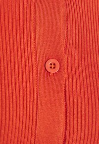 Červený žebrovaný pletený cardigan s kulatým čtyřdírkovým knoflíkem ve stejné barvě a jemnou texturou ze vzoru tkaniny.