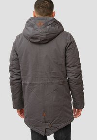 Grijze hooded parka met een gestructureerde afwerking, knoopaccenten en een klein bruin logopatch op de achterkant. Heeft een taps toelopende zoom en naden details.