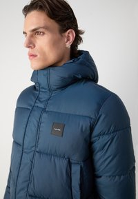 Giubbotto puffer blu navy con colletto alto, texture a costine, cuciture in evidenza e un'etichetta quadrata nera sul davanti.