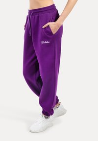 Fialové tepláky s vroubkovaným pasem a manžetami, se bočními kapsami a bílou vyšívanou logem na levém bok.