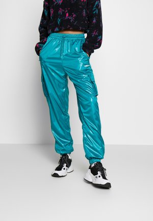 Persona con pantalones cargo Adidas color teal brillante con puños elásticos, combinados con zapatillas negras y blancas y una sudadera corta con capucha y patrón oscuro.