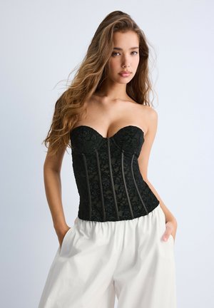 Jeune femme aux longs cheveux ondulés portant un corset en dentelle noire sans bretelles et un pantalon blanc ample, posant les mains dans les poches sur un fond uni.