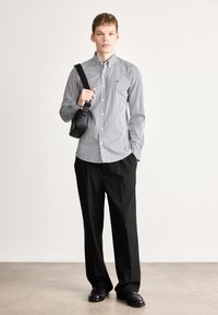 Ριγέ πουκάμισο με κουμπιά σε μαύρο και άσπρο, συνδυασμένο με μαύρο παντελόνι. Το μοντέλο κρατά μια μικρή μαύρη τσάντα χιαστί. Ομαλή υφή υφάσματος.