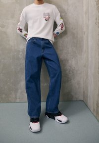 Camisa blanca de manga larga con gráficos de manos coloridos, combinada con jeans de corte relajado azul y zapatillas negras y blancas. Fondo de pared de hormigón.