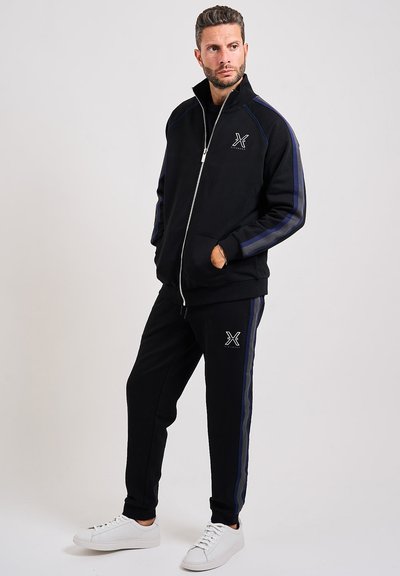 Tuta nera con zip e strisce blu a contrasto. Presenta un alto colletto, design slim-fit e dettagli con logo sulla giacca e sui pantaloni. Sneakers bianche.