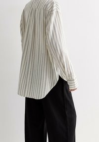 Langærmet button-up skjorte i creme med navy striber, lavet af letvægtsstof. Har en buet kant og en afslappet pasform. Kombineret med sorte bukser.
