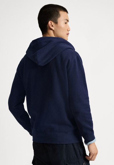 Mladenič, ki je obrnjen vstran, nosi mornarsko modro jopico s kapuco in črne hlače cargo na belem ozadju.