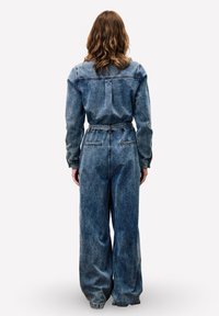Lichte denim jumpsuit met een relaxte pasvorm, elastische tailleband, lange mouwen en grote zakken; vervaagde textuur voegt visuele interesse toe.