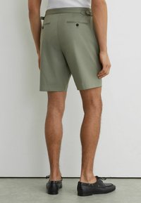 Hombre con pantalones cortos a medida de color verde claro con bolsillos traseros abotonados y zapatos negros de cuero con cordones, de pie contra una pared blanca.