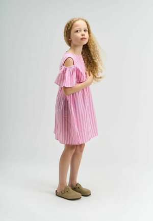 Noor tüdruk, kellel on pikad lokkis juuksed ja kellel on selja lahtise roosa triibulise kleidi ja beežide libisemisega kingadega, seistes valgel taustal.