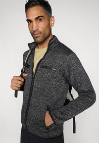 Ζακέτα φλις με φερμουάρ σε σκούρο γκρι με υφή, που διαθέτει τσέπη στο στήθος και πλαϊνές τσέπες με φερμουάρ, φορεμένη πάνω από ένα ανοιχτόχρωμο πουκάμισο.