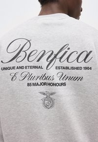 Άτομο που φορά γκρι φούτερ με τα κείμενα και το έμβλημα «Benfica», «Μοναδικό και Αιώνιο», «Ιδρύθηκε το 1904», «E Pluribus Unum» και «85 Μεγάλα Βραβεία».