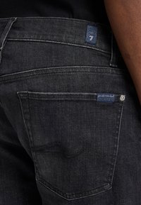 Donkerdenim jeans met een close-up van de achterzak, zilverkleurige hardwaredetails en een blauwe merklabel nabij de tailleband.