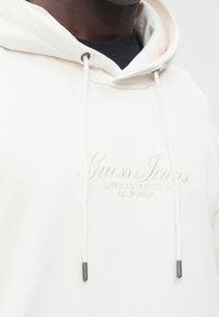 Sweat à capuche blanc en tissu doux, arborant un logo "Guess Jeans" brodé en ton sur ton, avec des cordons de serrage et des accents métalliques.