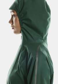 Roheline veekindel jope, millel on sobiv kapuuts, sile tekstuur, peegeldavad detailid ja voolujooneline siluett.