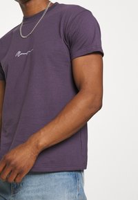 Fialové bavlněné tričko s krátkým rukávem, na přední straně je bílé nápisové logo. Kombinováno se světlými modrými džíny.