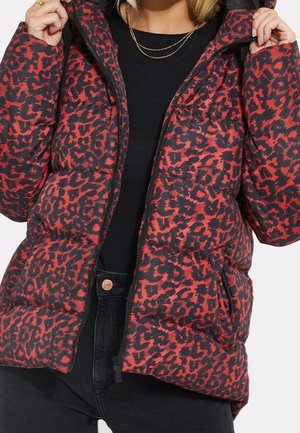 Kobieta w czerwonej i czarnej kurtce puchowej w panterkę, nosząca czarny top i dżinsy, trzymająca kołnierz kurtki obiema rękami.