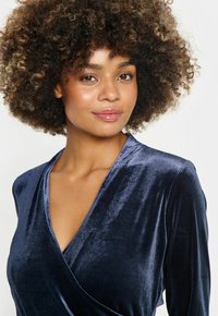 Navy blue velvet dress with a deep V-wrap neckline, long sleeves, and a soft, smooth texture. Features a subtle sheen and elegant draping.